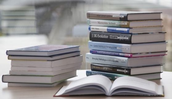 Two piles of books on a desk with an open book in the foreground.