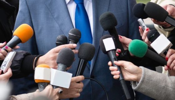 Close-up of journalists holding out mics to a man in a suit.
