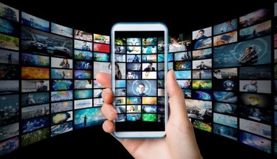 A hand holds up a mobile phone in front of a bank of screens.